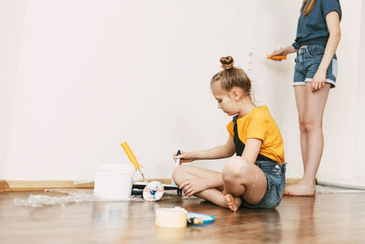 A girl with her sister in bright blue and yellow clothes helps to paint the walls in her room white.