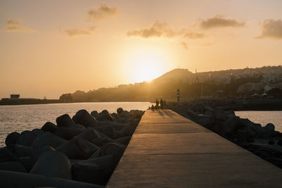 People on rocks by sea against sky during sunset