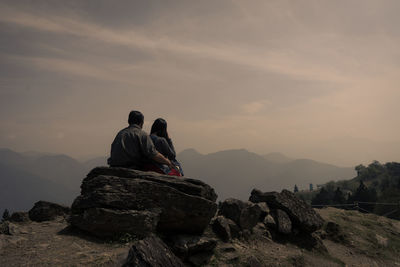 Rear view of couple sitting on rock against sky on sunny day