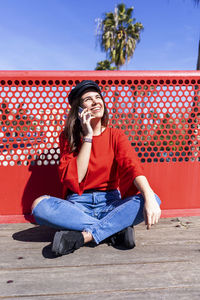 Teenage girl using mobile phone while wearing red top in city against railing during sunny day