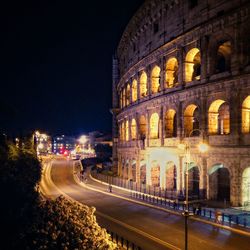 Illuminated coliseum by empty road in city at night