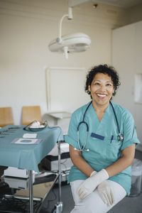 Portrait of happy female doctor wearing medical scrubs sitting in medical examination room