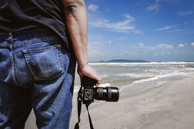Midsection of man with camera standing at beach against sky