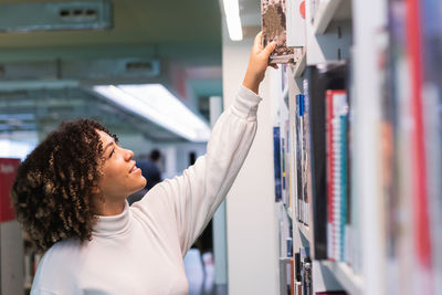 Side view of smiling african american female student taking book from shelf in library at university