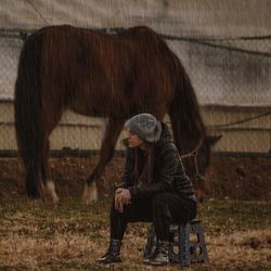 Full length of woman sitting on field beside a horse while raining