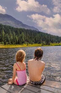 Rear view of siblings sitting on pier over lake against cloudy sky