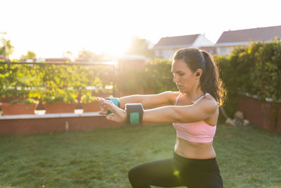 Side view of fit female athlete in sportswear using laptop and doing squats while following online tutorial during training in backyard at sunset