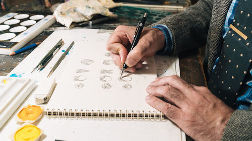 Side view of focused creative mature male designer working on jewelry designs and drawing sketches while sitting at table with professional supplies in workplace