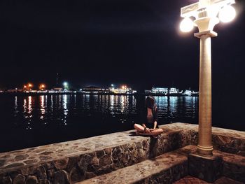 Woman sitting on retaining wall against sea at night