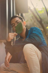 Portrait of young man holding leaf over mouth while crouching outdoors