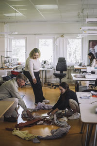 Female fashion designer assisting colleagues sorting recycled clothes on floor at workshop