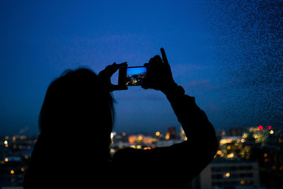 Silhouette woman photographing illuminated city against sky at night