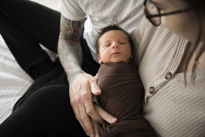Newborn boy swaddled in brown, held by tattooed hipster parents