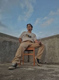 Portrait of young man sitting on bench against sky