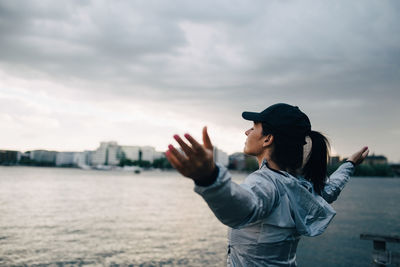 Female athlete standing with arms outstretched by sea against sky in city