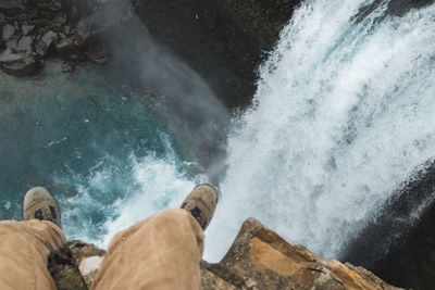 Low section of man sitting on cliff against waterfall