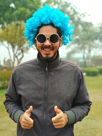 Portrait of smiling young man wearing sunglasses and blue wig standing at public park