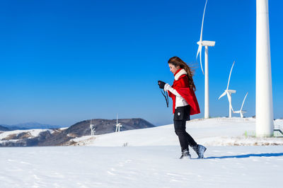 Woman holding camera while walking on snow covered road against wind turbines and clear blue sky