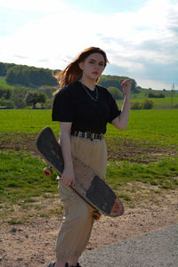 Young girl with a skateboard in her hand, stands in green nature, one arm up