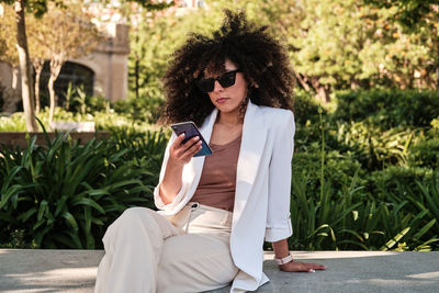 Hispanic female entrepreneur in white suit with curly hair browsing on smartphone while sitting on bench near fence on sunny day on city street