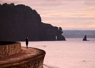 Rear view of woman jogging by sea against sky during sunset