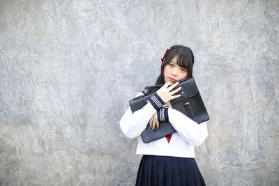 Portrait of young woman holding bag standing against wall