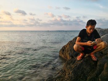 Young man using mobile phone while crouching at rocky beach during sunset