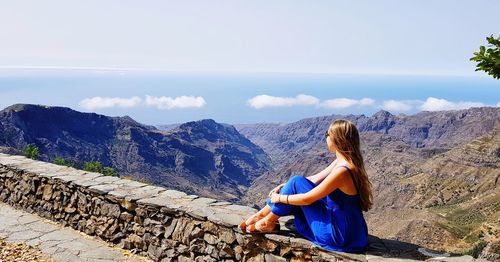 Woman sitting on retaining wall looking at mountains and sea during sunny day