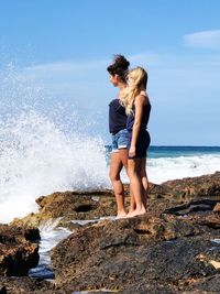 Full length of mother and daughter standing on rock at beach against sky