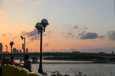 People on street by river against sky during sunset
