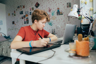 A teenage girl is studying in a room using a laptop and a drawing tablet.