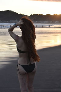 Rear view of woman in bikini standing at beach against sky during sunset