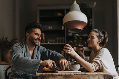 Happy non-binary person sharing smart phone with boyfriend while having breakfast at home