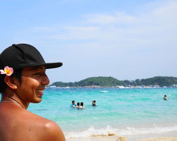 Rear view of happy shirtless man with frangipani and cap at beach against sky