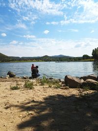 Rear view of man sitting on rock by lake against sky