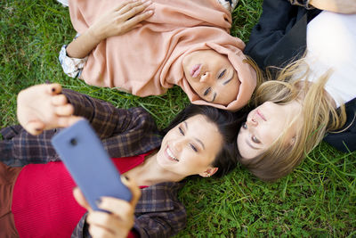 Smiling woman taking selfie with friends through smart phone lying on grass