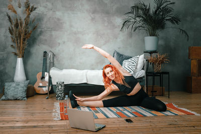 Positive athletic woman with red hair and tight sportswear stretching at home.