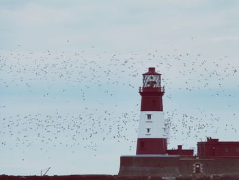 Flock of birds flying over lighthouse against sky
