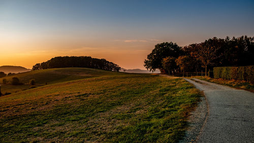 Road by trees on field against sky at sunset