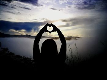 Woman hand holding heart shape against sky during sunset