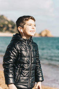 Smiling boy listening music while standing at beach against sky
