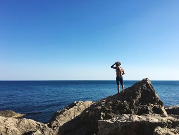 Rear view of shirtless man standing on rock at beach against clear blue sky during sunny day