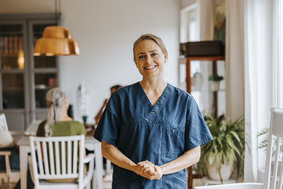 Portrait of smiling female nurse standing with hands clasped at care home