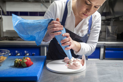 Female chef in apron using pastry bag to squeeze sweet cream on plate while cooking dessert on metal table in restaurant kitchen