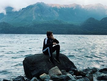 Boy sitting on rock by lake against mountain