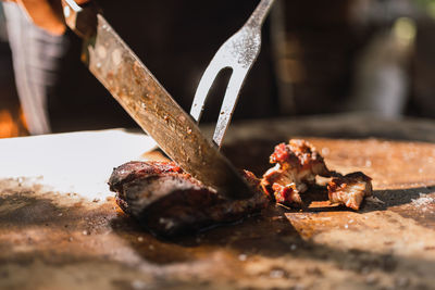 Cropped unrecognizable person with knife and carving fork cutting yummy meat piece on board against barbecue rack in sunlight person