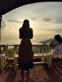 Rear view of couple standing on shore against sunset sky