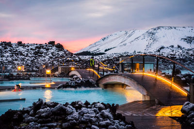 Bridge over river by snowcapped mountains against sky during sunset