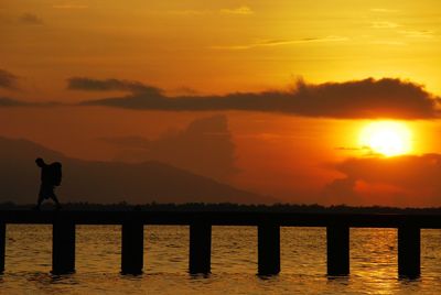Silhouette traveler walking on pier over sea against orange sky