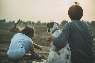 Rear view of mother and daughter sitting with dog on field against clear sky during sunset